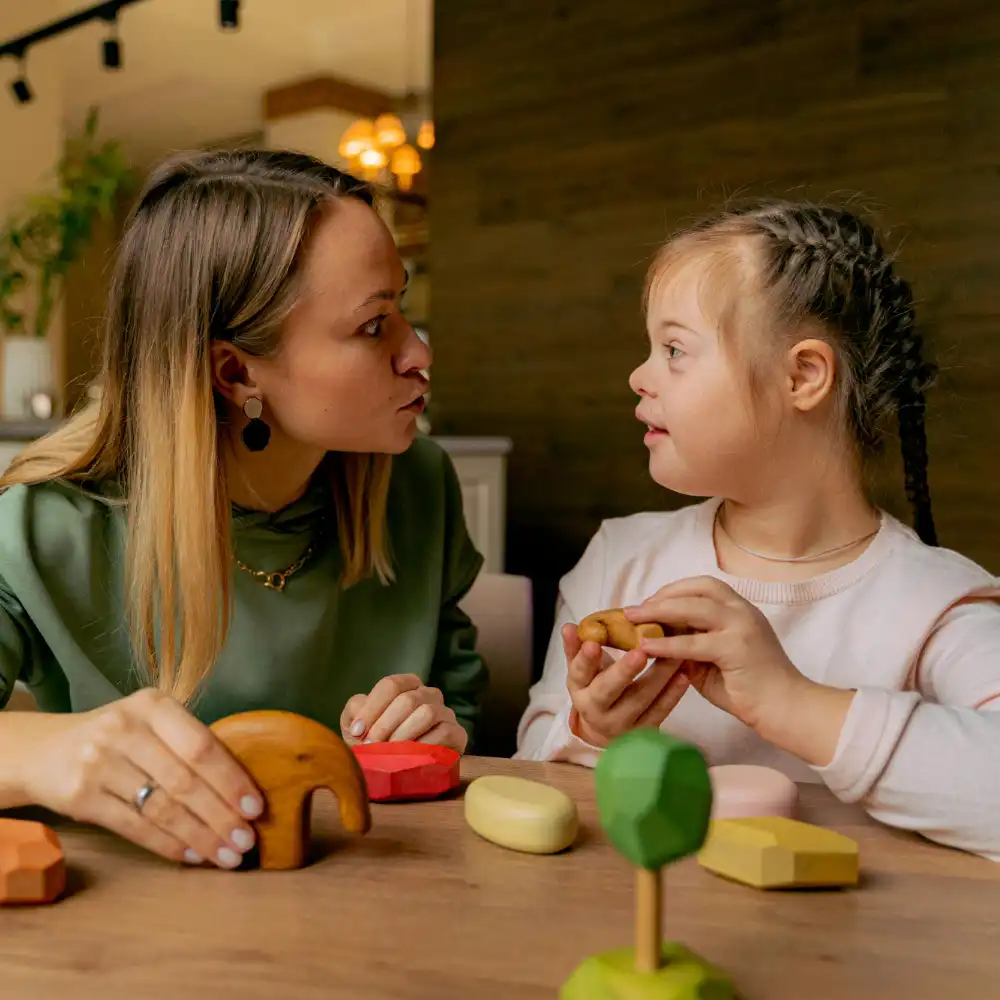 Teacher providing one-on-one learning support to a young girl with Down syndrome using educational wooden toys at a table.