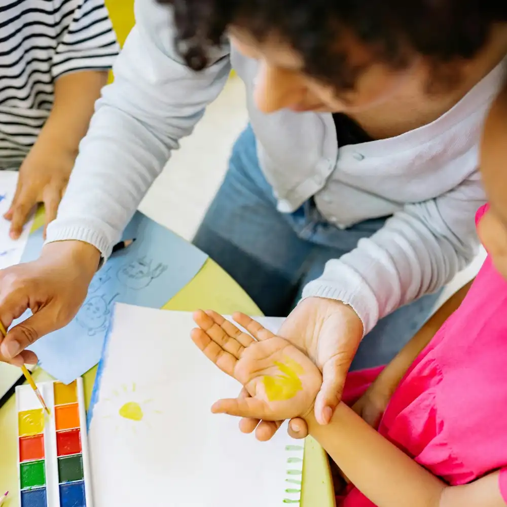 Teacher guiding a young child in a watercolor painting activity at a table with art supplies.